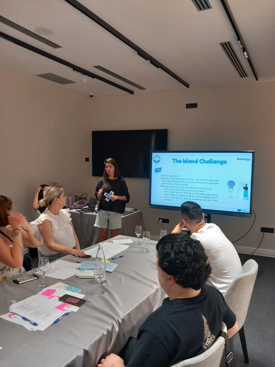 Trener vodi radionicu, predstavlja sadržaj na ekranu, dok učesnici sjede oko stola s bilješkama i materijalima u sali za sastanke.