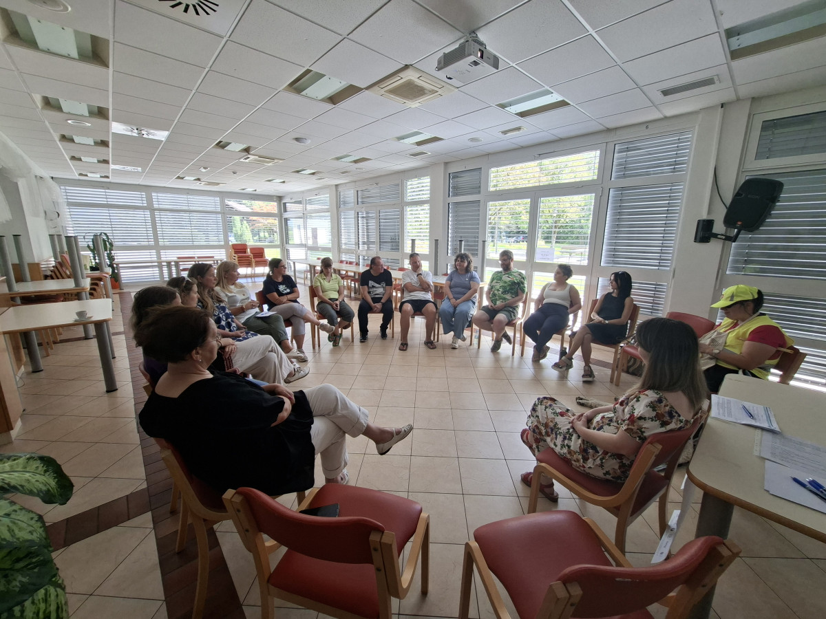 Grupa učesnika iz Slovenije sjedi u krugu na stolicama u svijetlom prostoru i učestvuje u grupnoj raspravi.