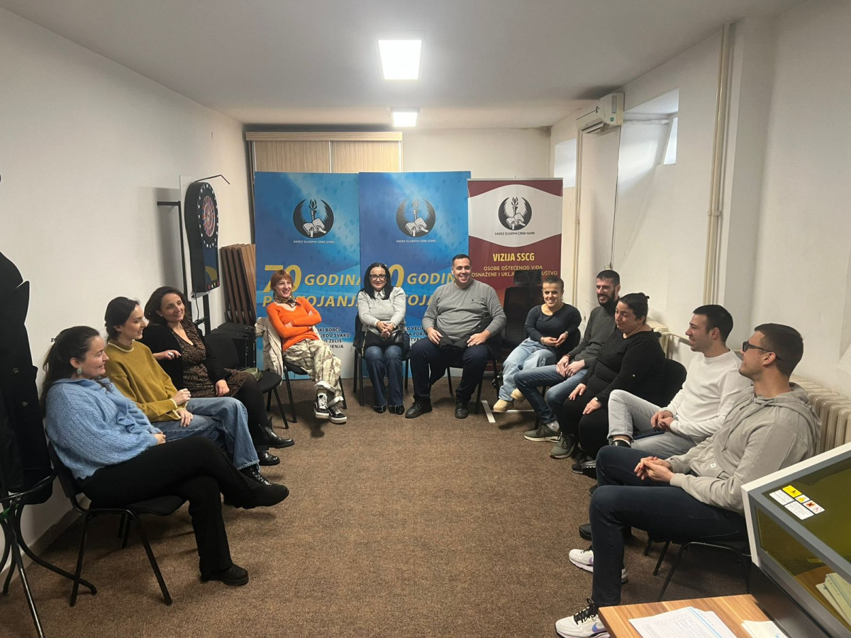 A group of fifteen people from Montenegro seated in a circle in a well-lit room. Two banners are visible in the background, featuring text in blue and burgundy. Participants include men and women of various ages, engaged in conversation.