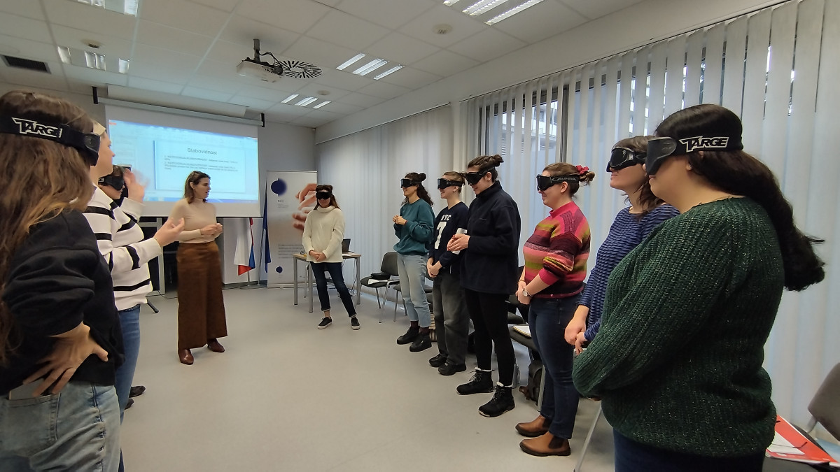 A group of participants from Slovenia wearing blindfolds stands in a classroom setting. A woman in a beige top gestures as she speaks, while others face her in various colored clothing. Bright windows and a presentation screen are visible in the background.