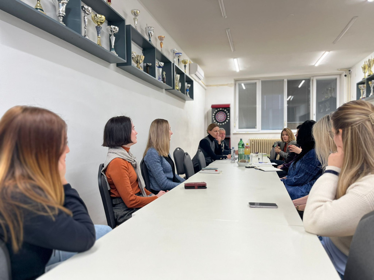 A group of women from Croatia sit around a long white table in a well-lit room, engaged in discussion. The wall displays various trophies on shelves above. A dartboard is visible in the background, and some participants face each other while others look away.
