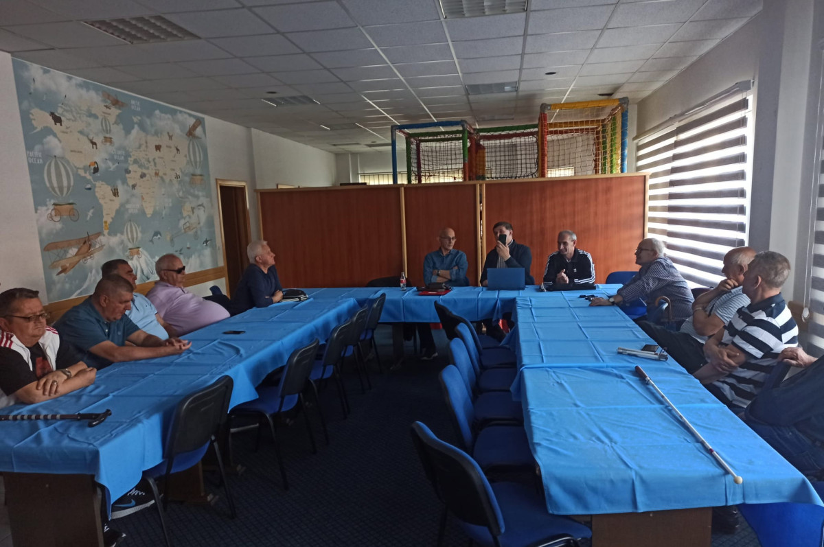 A group of men from Bosnia and Herzegovina are seated around long blue tables in a well-lit meeting room. They are engaged in discussion, with some looking at each other and others at a speaker. The room features a mural on one wall and a play area in the background.