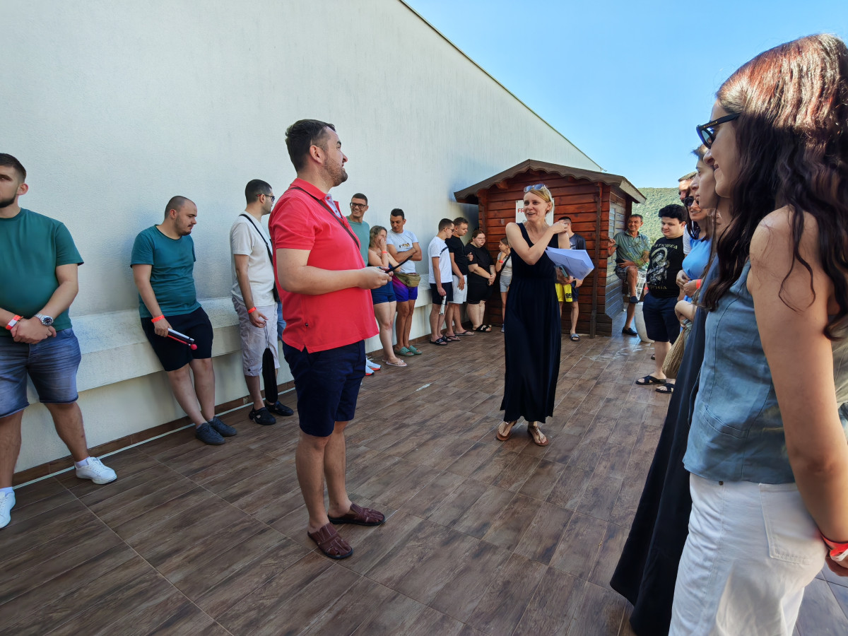 Μια ομάδα συμμετεχόντων από όλες τις χώρες των εταίρων, μεταξύ των οποίων αρκετά άτομα που χρησιμοποιούν λευκά μπαστούνια και φορούν γυαλιά, ακούει μια εξήγηση σε μια ηλιόλουστη βεράντα με ξύλινο δάπεδο. Στα αριστερά, ένας άνδρας συμμετέχων από τη Βοσνία και Ερζεγοβίνη, που φοράει κόκκινη κοντομάνικη μπλούζα, μιλάει. Στο κέντρο, μια εισηγήτρια με μακρύ μαύρο φόρεμα συνομιλεί με τον άνδρα με την κόκκινη μπλούζα, κρατώντας έγγραφα στο χέρι της. Άλλοι συμμετέχοντες στέκονται στο βάθος, κοντά σε έναν ψηλό λευκό τοίχο και ένα ξύλινο κτίριο με μια πινακίδα που φέρει έναν κόκκινο σταυρό. Όλοι είναι ντυμένοι με casual ρούχα.
