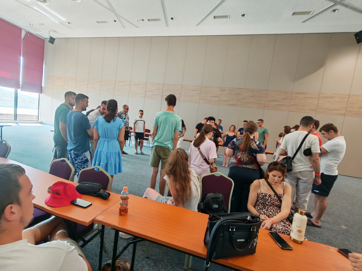 Μια μεγαλύτερη ομάδα συμμετεχόντων στέκεται σε κύκλους ή μικρότερες ομάδες κατά τη διάρκεια μιας δραστηριότητας στο πλαίσιο ενός εργαστηρίου, σε μια ευρύχωρη αίθουσα. Στο προσκήνιο υπάρχουν μακριά τραπέζια με προσωπικά αντικείμενα, όπως τσάντες, ποτά και ένα κόκκινο καπέλο. Η αίθουσα έχει ψηλή οροφή, γκρι χαλί και μεγάλα παράθυρα στην αριστερή πλευρά, από τα οποία εισέρχεται φυσικό φως.