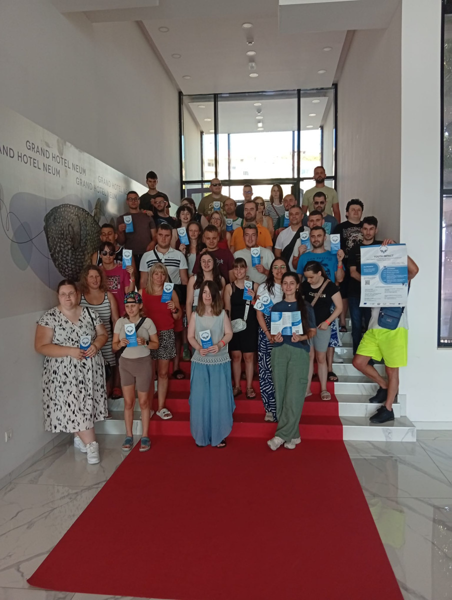 Ολόκληρη η ομάδα των συμμετεχόντων, των εθελοντών και των επικεφαλής των εθνικών ομάδων που λαμβάνουν μέρος στην εκπαίδευση νέων ποζάρει σε μια σκάλα καλυμμένη με κόκκινο χαλί, στο λόμπι του Grand Hotel Neum. Αρκετά άτομα κρατούν μπλε φυλλάδια ή ενημερωτικά έντυπα του έργου, ενώ ένας εθελοντής στα δεξιά κρατά μια μεγαλύτερη αφίσα με τις λέξεις «Youth Impact». Ο χώρος είναι φωτεινός, με μεγάλες γυάλινες πόρτες στο βάθος και την ονομασία του ξενοδοχείου να εμφανίζεται στον τοίχο στα αριστερά.